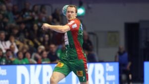 Ein männlicher Handballspieler in einem grün-roten Trikot macht sich während eines Spiels bereit, den Ball zu werfen; im Hintergrund sind das Publikum und die Anzeigetafel unscharf zu sehen.