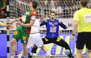 Ein Handballtorwart in Blau macht sich bereit, einen Wurf abzuwehren, während ein gegnerischer Spieler in Grün zum Wurf ansetzt. Auf dem Spielfeld sind außerdem ein weiterer Spieler in Weiß und ein Schiedsrichter in Gelb zu sehen. Im Hintergrund verfolgt das Publikum das Geschehen.