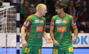 Zwei männliche Handballspieler in grünen und roten Trikots stehen nebeneinander auf dem Spielfeld und scheinen sich über etwas zu unterhalten. Einer trägt eine Brille; beide haben Sponsorenlogos auf ihren Trikots. Im Hintergrund ist ein Tor zu sehen.