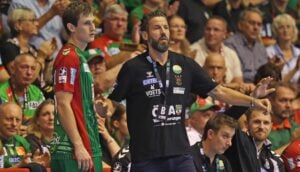 Ein Handballtrainer gestikuliert während eines Spiels neben einem Spieler in einem grün-roten Trikot, während im Hintergrund eine Menge Zuschauer zuschaut.