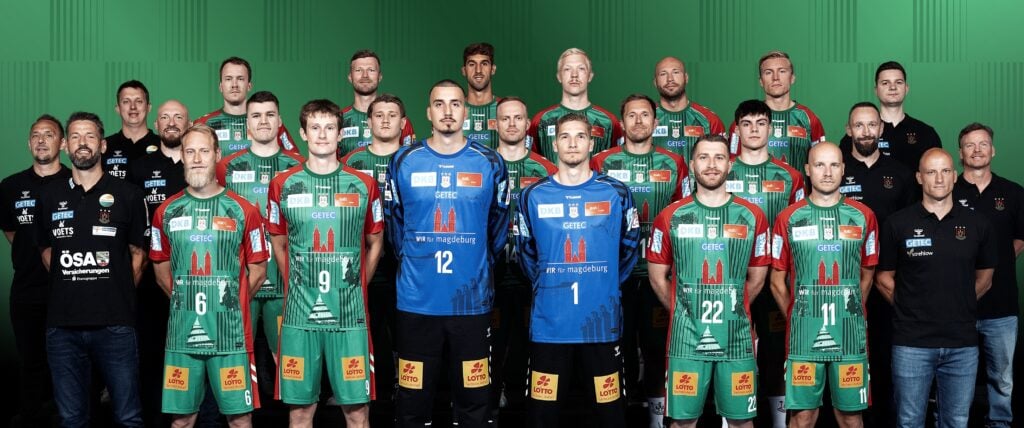 45_vor_SCM_Teambild Eine Herren-Handballmannschaft posiert in grünen und roten Trikots zusammen mit Trainern und Betreuern in schwarzen Hemden und steht in drei Reihen vor einem grünen Hintergrund.