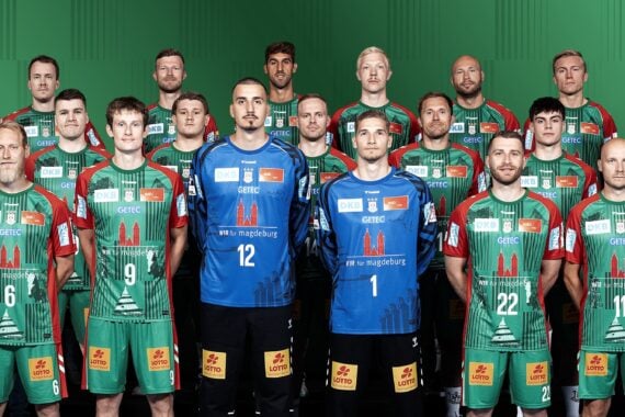 Eine Herren-Handballmannschaft posiert in grünen und roten Trikots zusammen mit Trainern und Betreuern in schwarzen Hemden und steht in drei Reihen vor einem grünen Hintergrund.