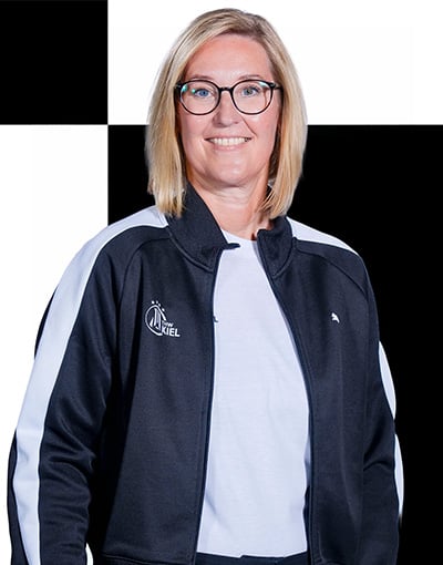 Eine lächelnde Frau mit blonden Haaren und Brille, möglicherweise eines der Mitglieder des Aufsichtsrats des THW Kiel, trägt eine schwarz-weiße Sportjacke über einem weißen Hemd und steht vor einem schwarz-weiß karierten Hintergrund.