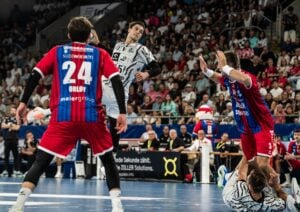 Ein Handballspieler in weißer Spielkleidung springt zum Wurf ab, während zwei Spieler in roten Trikots verteidigen. Die Zuschauer im Hintergrund verfolgen diesen spannenden Moment des Spiels.