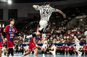 Ein Handballspieler in einem schwarz-weißen Trikot springt in der Luft, um den Ball zu werfen, während Gegner in roten Trikots zusehen. Die Szene spielt sich in einer vollbesetzten Halle mit Zuschauern ab.