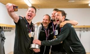 Vier Männer in einheitlichen Mannschaftstrikots feiern mit Medaillen und einem großen Pokal, lächeln und machen gemeinsam ein Selfie in einer Umkleidekabine.