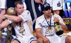 Zwei männliche Sportler in weißen Trikots sitzen auf dem Boden und feiern. Der eine umarmt einen großen Pokal, während der andere eine Champagnerflasche in der Hand hält und eine schwarze Mütze trägt. Um sie herum liegt Konfetti verstreut.