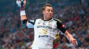 Ein Handballspieler in einem schwarz-weißen Trikot springt in die Höhe und macht sich bereit, den Ball zu werfen, während im Hintergrund ein vollbesetztes Stadion zu sehen ist.