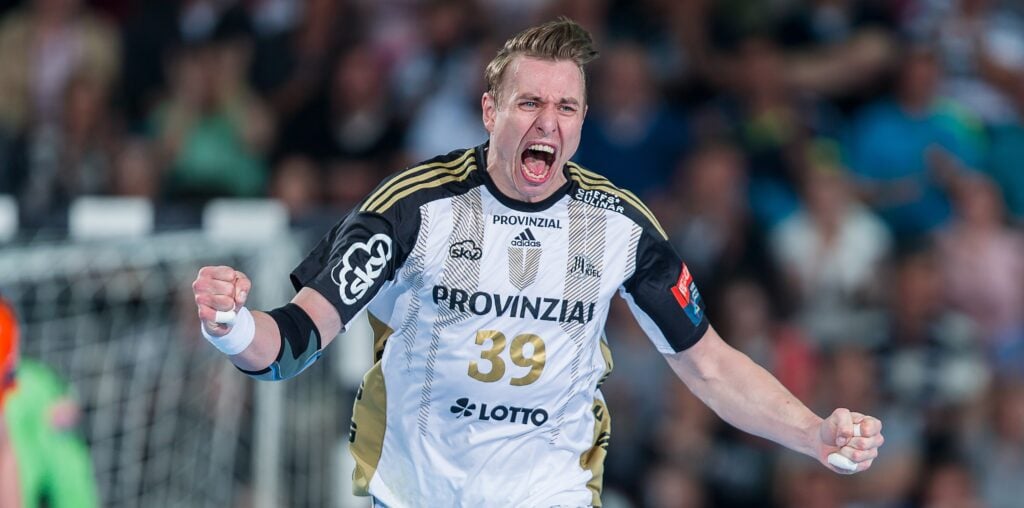 Ein Handballspieler in einem schwarz-weißen Trikot mit der Nummer 39 feiert während eines Spiels leidenschaftlich mit geballten Fäusten, im Hintergrund ist eine verschwommene Menschenmenge zu sehen.