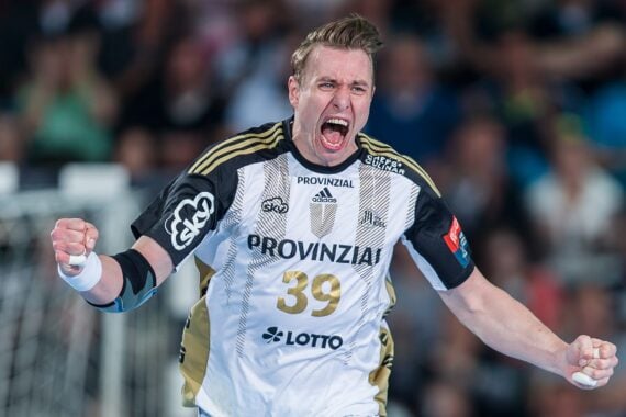 Ein Handballspieler in einem schwarz-weißen Trikot mit der Nummer 39 feiert während eines Spiels leidenschaftlich mit geballten Fäusten, im Hintergrund ist eine verschwommene Menschenmenge zu sehen.