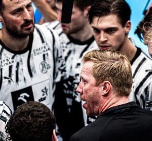 Ein Trainer spricht während eines Spiels leidenschaftlich mit seiner Mannschaft, die schwarz-weiße Trikots trägt. Die Spieler hören aufmerksam zu und blicken dabei konzentriert und ernst.