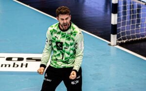 Ein Handballtorwart in einem grünen Trikot jubelt begeistert, ballt beide Fäuste und schreit vor dem Tor in einer Halle.