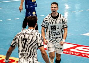 Zwei männliche Handballspieler in schwarz-weiß gestreiften Trikots feiern auf einem Hallenfeld. Der eine jubelt mit vor Aufregung geballten Fäusten, während der andere ihm gegenübersteht. Im Hintergrund sind das blaue Spielfeld und Werbeplakate zu sehen.