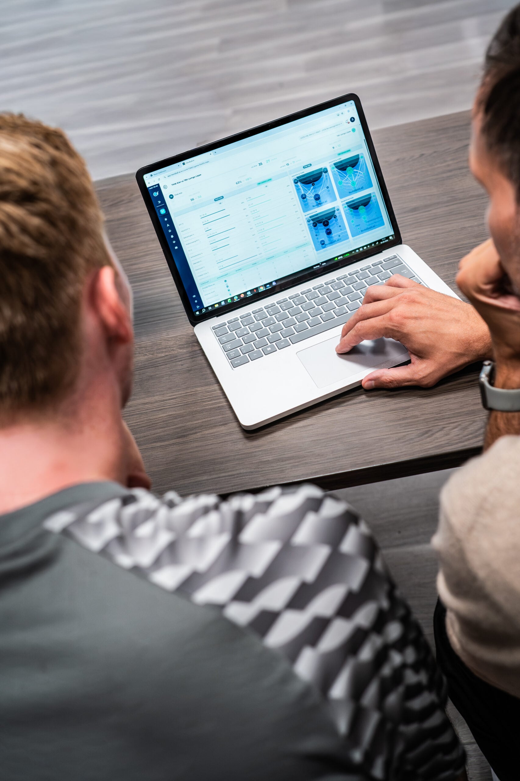 Zwei Personen sitzen an einem Tisch und schauen auf einen Laptop, auf dessen Bildschirm Grafiken und Diagramme zu sehen sind. Während sie gemeinsam die Daten durchgehen – und dabei vielleicht über aktuelle Entwicklungen sprechen, wie zum Beispiel: „Die zweite Runde im DHB-Pokal wurde ausgelost. Das ist der Gegner des THW Kiel.“ –, navigiert eine der beiden Personen mithilfe des Touchpads des Laptops durch die Informationen.