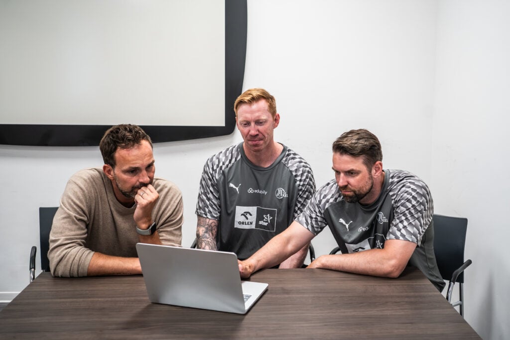 Handball.ai-3 Drei Männer sitzen an einem Tisch in einem Besprechungsraum und schauen gebannt auf den Bildschirm eines Laptops. Zwei von ihnen tragen identische graue Sporthemden, während der dritte einen hellbraunen Pullover trägt. An der Wand hinter ihnen hängt eine große leere Leinwand. Vielleicht schauen sie sich gerade an, wie die zweite Runde im DHB-Pokal ausgelost wurde und wer der Gegner des THW Kiel ist.