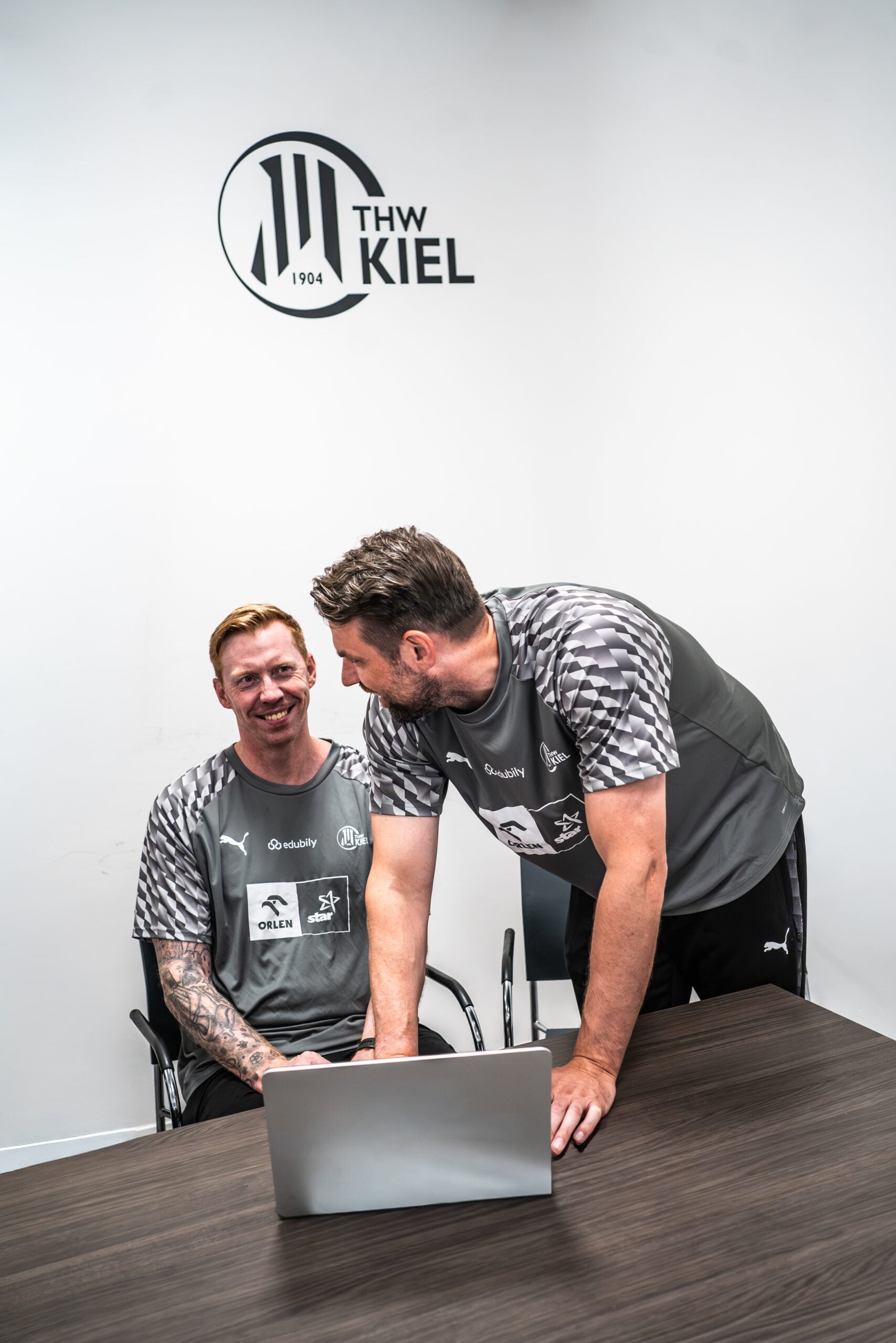 Zwei Männer in THW-Kiel-Sporttrikots sitzen und stehen an einem Tisch neben einem Laptop, lächeln und unterhalten sich, während hinter ihnen das THW-Kiel-Logo an der weißen Wand zu sehen ist. Die zweite Runde im DHB-Pokal wurde ausgelost – das ist der Gegner des THW Kiel, über den die beiden offenbar diskutieren.