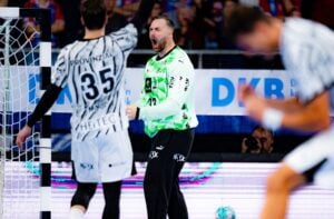 Ein Handballtorwart in einem neongrünen Trikot feiert einen erfolgreichen Spielzug, ballt die Faust und jubelt, während seine Teamkollegen in schwarz-weißen Trikots neben ihm auf dem Spielfeld reagieren.