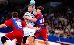 Ein Handballspieler in weißer Spielkleidung springt zum Wurf ab, während zwei Gegner in roten Trikots ihn während eines spannenden Spiels in einer vollbesetzten Halle verteidigen.