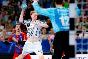 Ein Handballspieler in einem schwarz-weißen Trikot springt hoch, um den Ball zu werfen, und zielt dabei am Torwart in Blau vorbei. Andere Spieler und Zuschauer verfolgen das Geschehen gebannt.