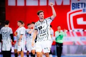 Ein Handballspieler in einem schwarz-weißen Trikot reckt jubelnd die Faust auf dem Spielfeld in die Höhe, im Hintergrund sind seine Mannschaftskameraden und ein großes rotes Transparent zu sehen.