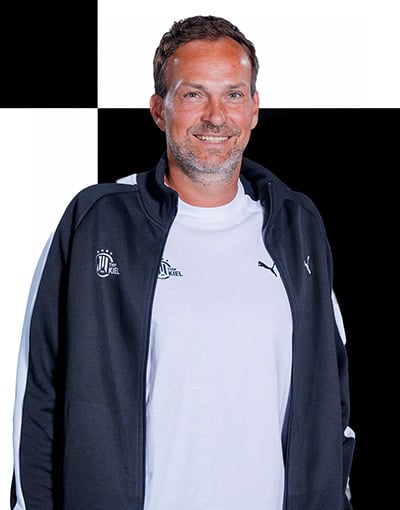 Ein Mann mit kurzen braunen Haaren und Bart, der ein weißes Hemd und eine schwarz-weiße Jacke mit Reißverschluss und Sportlogos trägt, steht lächelnd vor einem schwarz-weiß karierten Hintergrund – ein Symbol für die Professionalität, die man häufig bei den Mitgliedern der Geschäftsführung und des Wirtschaftsausschusses des THW Kiel antrifft.