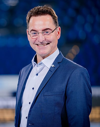 Ein Mann in einem blauen Sakko und mit Brille lächelt in die Kamera, während er vor einem unscharfen blauen Hintergrund steht – ein Sinnbild für die Professionalität, die man oft bei den Mitgliedern der Geschäftsführung und den Vertretern des Aufsichtsrats des THW Kiel sieht.