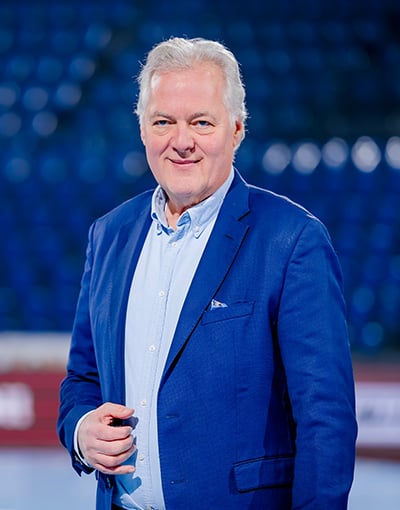 Ein Mann mit grauen Haaren, der einen blauen Anzug und ein hellblaues Hemd trägt, steht vor verschwommenen blauen Stadionsitzen, lächelt in die Kamera und verkörpert die Professionalität, die man oft bei Mitgliedern der Geschäftsführung oder des Aufsichtsrats des THW Kiel sieht.