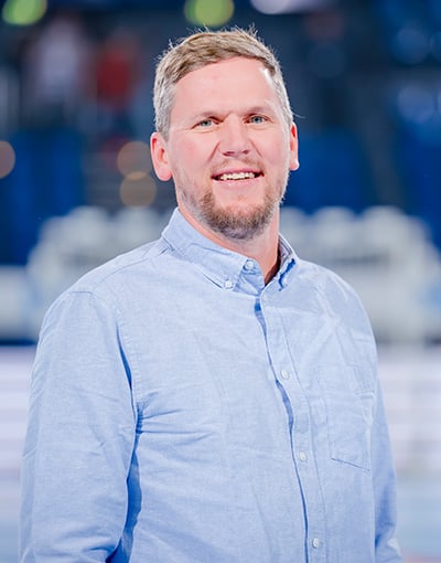 Ein Mann mit kurzen hellbraunen Haaren und Bart, der ein hellblaues Hemd mit Knöpfen trägt, lächelt, während er in einem Raum vor einem verschwommenen blau-weißen Hintergrund steht – er ist eines der engagierten Mitglieder im THW Kiel-Wirtschaftsausschuss.