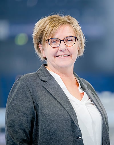 Eine Frau mit kurzen blonden Haaren und Brille lächelt; sie trägt einen grauen Blazer über einer weißen Bluse und steht vor einem verschwommenen blau-grauen Hintergrund – ein Bild, das zu einem Mitglied der Geschäftsführung des THW Kiel oder zu einem Mitglied des Aufsichtsrats des Vereins passen würde.
