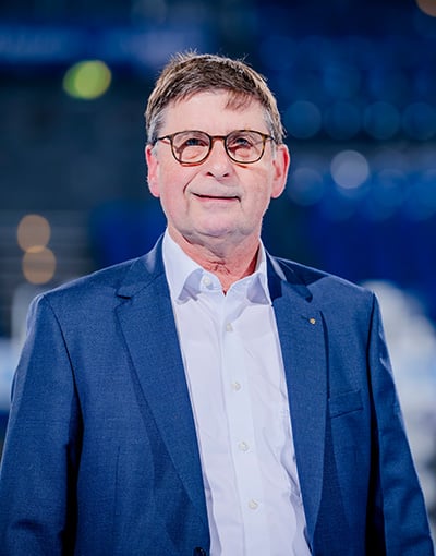 Ein Mann mit Brille, weißem Hemd und blauem Blazer steht in einem Raum und blickt in die Kamera. Der unscharfe Hintergrund ist geprägt von Blautönen und Lichtreflexen, was die professionelle Atmosphäre vermittelt, die oft mit Sitzungen der THW Kiel-Geschäftsführung oder der Mitglieder des Wirtschaftsausschusses in Verbindung gebracht wird.