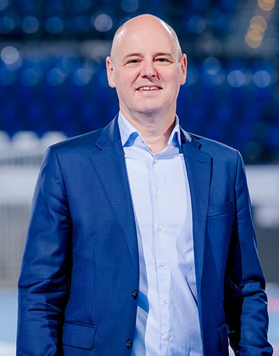 Ein lächelnder, kahlköpfiger Mann in einem blauen Sakko und einem hellblauen Hemd steht in einem Raum, im Hintergrund sind verschwommene blaue Sitzplätze zu sehen – ein Symbol für die Professionalität, die man oft bei den Mitgliedern der THW Kiel-Geschäftsführung beobachtet.