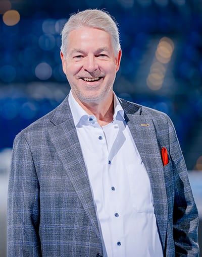 Ein lächelnder Mann mit grauen Haaren, der einen grauen karierten Blazer, ein weißes Hemd und ein rotes Einstecktuch trägt, steht vor einem unscharfen blauen Hintergrund – ideal, um wichtige Persönlichkeiten aus der Geschäftsführung des THW Kiel oder Mitglieder des Aufsichtsrats hervorzuheben.