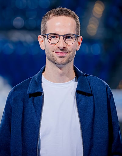 Ein Mann mit kurzen braunen Haaren und Brille, der ein weißes T-Shirt und eine blaue Jacke trägt, lächelt in die Kamera. Der Hintergrund ist verschwommen und weist blaue und weiße Farbtöne sowie kreisförmige Lichter auf, was darauf hindeutet, dass er möglicherweise zu den Mitgliedern der Geschäftsführung des THW Kiel oder zum Aufsichtsrat gehört.