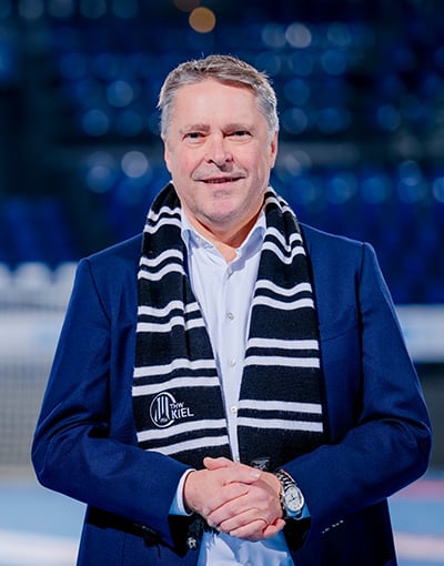 Ein Mann in einem blauen Blazer und einem gestreiften Schal steht in einem Raum und lächelt in die Kamera. Der Hintergrund ist unscharf und zeigt blaue Stadionsitze – eine Umgebung, die daran erinnert, wo sich die Mitglieder der Geschäftsführung und des Aufsichtsrats des THW Kiel zu Sitzungen des Wirtschaftsausschusses versammeln könnten.