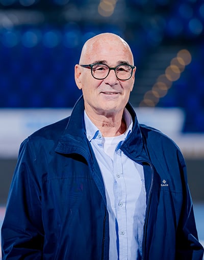 Ein kahlköpfiger Mann mit Brille, der ein hellblaues Hemd und eine dunkelblaue Jacke trägt, steht in einem Raum und lächelt sanft. Als eines der Mitglieder des Wirtschaftsausschusses strahlt er ruhiges Selbstvertrauen aus. Der Hintergrund ist unscharf, in Blautönen gehalten und von verschwommenen Lichtern geprägt.