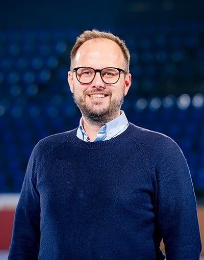 Vor einem verschwommenen blauen Stadionhintergrund steht ein lächelnder Mann mit Brille, kurzem Bart und hellbraunen Haaren, der einen marineblauen Pullover über einem Hemd mit Kragen trägt. Als eines der Mitglieder des Aufsichtsrats der Geschäftsführung des THW Kiel bringt er Fachwissen und Engagement in den Wirtschaftsausschuss ein.