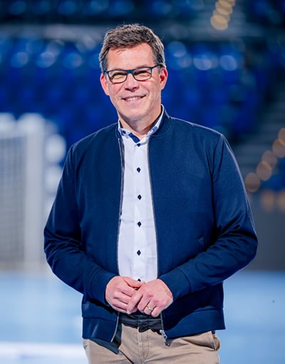 Ein lächelnder Mann mit kurzen braunen Haaren und Brille trägt eine marineblaue Jacke über einem weißen Hemd. Er steht in einem Raum mit blauen Stadion-Sitzen und verschwommenen Lichtern im Hintergrund und verkörpert die Professionalität, die man oft bei den Mitgliedern der THW Kiel-Geschäftsführung sieht.