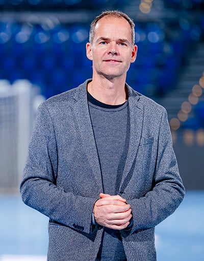 Ein Mann in grauem Blazer und Hemd steht in einem Innenraum und hat die Hände gefaltet. Der Hintergrund ist unscharf, doch man erkennt blaue Stadionsitze und ein Sportnetz, was auf eine Umgebung hindeutet, die der Geschäftsführung von THW Kiel oder vielleicht den Mitgliedern des Aufsichtsrats bei einer Sportveranstaltung vertraut ist.