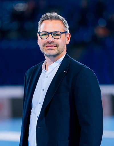 Ein Mann mit kurzen Haaren, Brille und gepflegtem Bart trägt einen dunklen Blazer über einem hellen Hemd und steht in einem Innenraum vor einem verschwommenen blauen Hintergrund – sein gelassenes Auftreten spiegelt die Professionalität wider, die man oft bei den Mitgliedern der Geschäftsführung und des Aufsichtsrats des THW Kiel sieht.