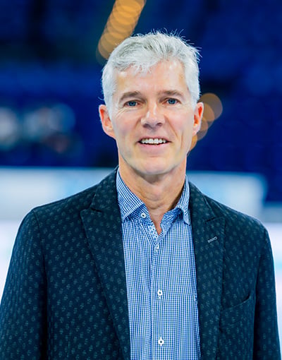 Ein Mann mittleren Alters mit grauen Haaren lächelt; er trägt einen blau gemusterten Blazer über einem blau karierten Hemd und steht in einem Innenraum vor einem verschwommenen blauen Hintergrund – ein Bild, das die professionelle Ausstrahlung widerspiegelt, die man oft bei Mitgliedern der Geschäftsführung oder des Aufsichtsrats des THW Kiel sieht.