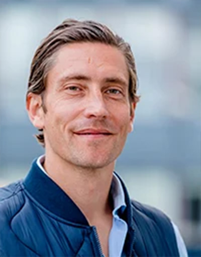 Ein Mann mit kurzen braunen Haaren, der eine blaue Jacke und ein hellblaues Hemd trägt – möglicherweise eines der Mitglieder der Geschäftsführung oder des Wirtschaftsausschusses des THW Kiel – lächelt leicht, während er im Freien steht; der Hintergrund ist unscharf.