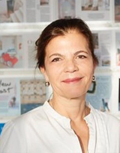 Eine Frau mit nach hinten gebundenem dunklem Haar, die eine weiße Bluse trägt und vor einer Wand steht, die mit verschiedenen Zeitschriftenseiten oder Artikeln bedeckt ist. Sie lächelt sanft und blickt in die Kamera, was die gelassene Professionalität vermittelt, die man oft bei den Mitgliedern der THW Kiel-Geschäftsführung sieht.