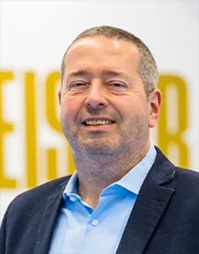 Ein Mann mit kurzen Haaren und Bart lächelt; er trägt ein blaues Hemd und einen dunklen Blazer. Der Hintergrund ist unscharf, wobei teilweise große gelbe Buchstaben zu erkennen sind, was darauf hindeutet, dass er eines der Mitglieder im Aufsichtsrat oder im Wirtschaftsausschuss der THW Kiel-Geschäftsführung sein könnte.