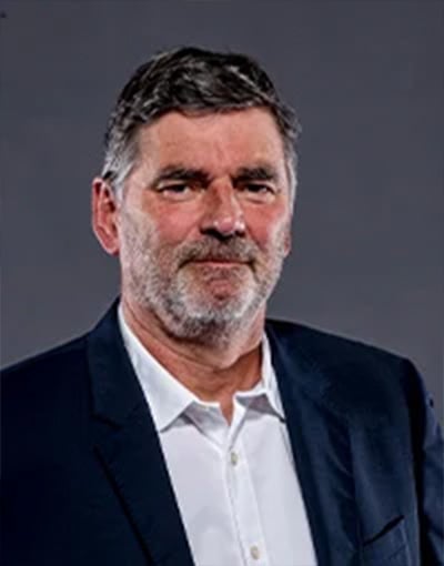 Ein Mann mittleren Alters mit kurzen grauen Haaren und Bart, der ein weißes Hemd und ein dunkles Sakko trägt, posiert vor einem schlichten grauen Hintergrund – ganz im Sinne der Professionalität, die von den Mitgliedern der THW Kiel-Geschäftsführung erwartet wird.