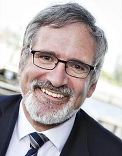 Ein lächelnder älterer Mann mit grauen Haaren, Bart und Brille, gekleidet in einen dunklen Anzug, ein weißes Hemd und eine gestreifte Krawatte – er strahlt Selbstbewusstsein und Professionalität aus. Der Hintergrund ist unscharf und zeigt eine Außenaufnahme. Er wirkt so, als könnte er zu den Mitgliedern der Geschäftsführung des THW Kiel gehören oder vielleicht Mitglied des Aufsichtsrats oder des Wirtschaftsausschusses sein.
