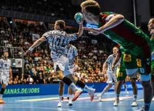 Ein Handballspieler in einem weißen Trikot springt während eines spannenden Hallenspiels in die Höhe, um den Ball zu werfen – eine Szene, die daran erinnert, wie der THW Kiel in der Handball-Bundesliga gegen den Titelverteidiger SC Magdeburg verloren hat.