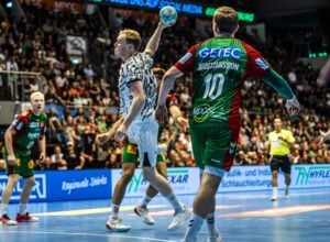 Ein Handballspieler in einem weiß gemusterten Trikot springt zum Wurf hoch, während Verteidiger in Grün und Rot ihn abdecken – so wie auch der THW Kiel in der Handball-Bundesliga beim Titelverteidiger SC Magdeburg verloren hat.