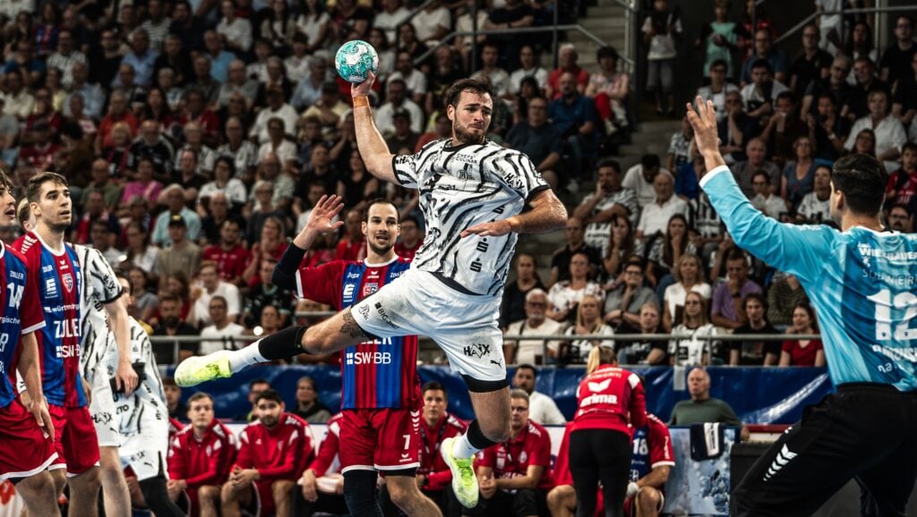 Ein Handballspieler in einem schwarz-weißen Trikot springt zum Wurf hoch und zielt auf das Tor, während der Torwart und die Spieler in Rot versuchen, ihn zu blocken. Die Zuschauer im Hintergrund verfolgen das Geschehen gespannt.