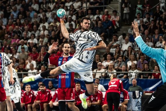 Ein Handballspieler in einem schwarz-weißen Trikot springt zum Wurf hoch und zielt auf das Tor, während der Torwart und die Spieler in Rot versuchen, ihn zu blocken. Die Zuschauer im Hintergrund verfolgen das Geschehen gespannt.