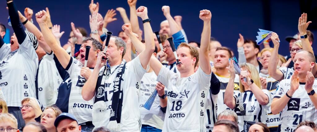 HANDBALL 2025/26: SG Flensburg-Handewitt - THW Kiel Eine Menge begeisterter Sportfans in einheitlichen weißen Trikots jubelt und reckt bei einer Hallenveranstaltung im Gleichklang die Fäuste in die Höhe – ein Bild, das die elektrisierende Atmosphäre des ausverkauften Derbys zwischen Kiel und Flensburg in der Handball-Bundesliga einfängt.